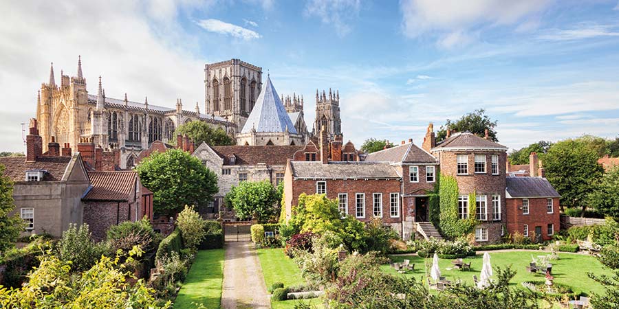 View of York Minster from the York city walls with blue skies. Overlooking gardens and trees in the foreground, with chairs and tables on the lawn. 