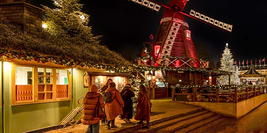 The Christmas markets of Copenhagen at night, tourists walk towards a festive windmill amongst Christmas lights and decorations. 